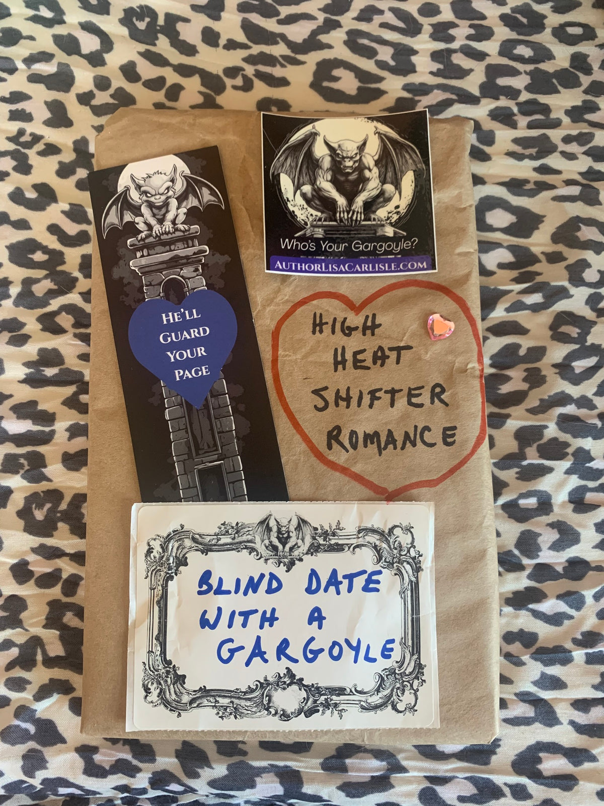 A brown paper mailer envelope decorated with three custom gargoyle-themed stickers and handwritten notes, placed on a leopard print fabric background.