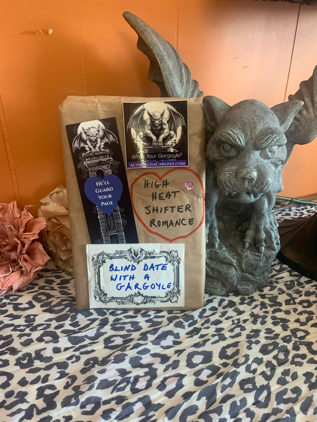 A brown paper bag package with multiple stickers and handwritten labels, standing on a leopard-print surface in front of an orange wall. A gray gargoyle figurine is beside the package.