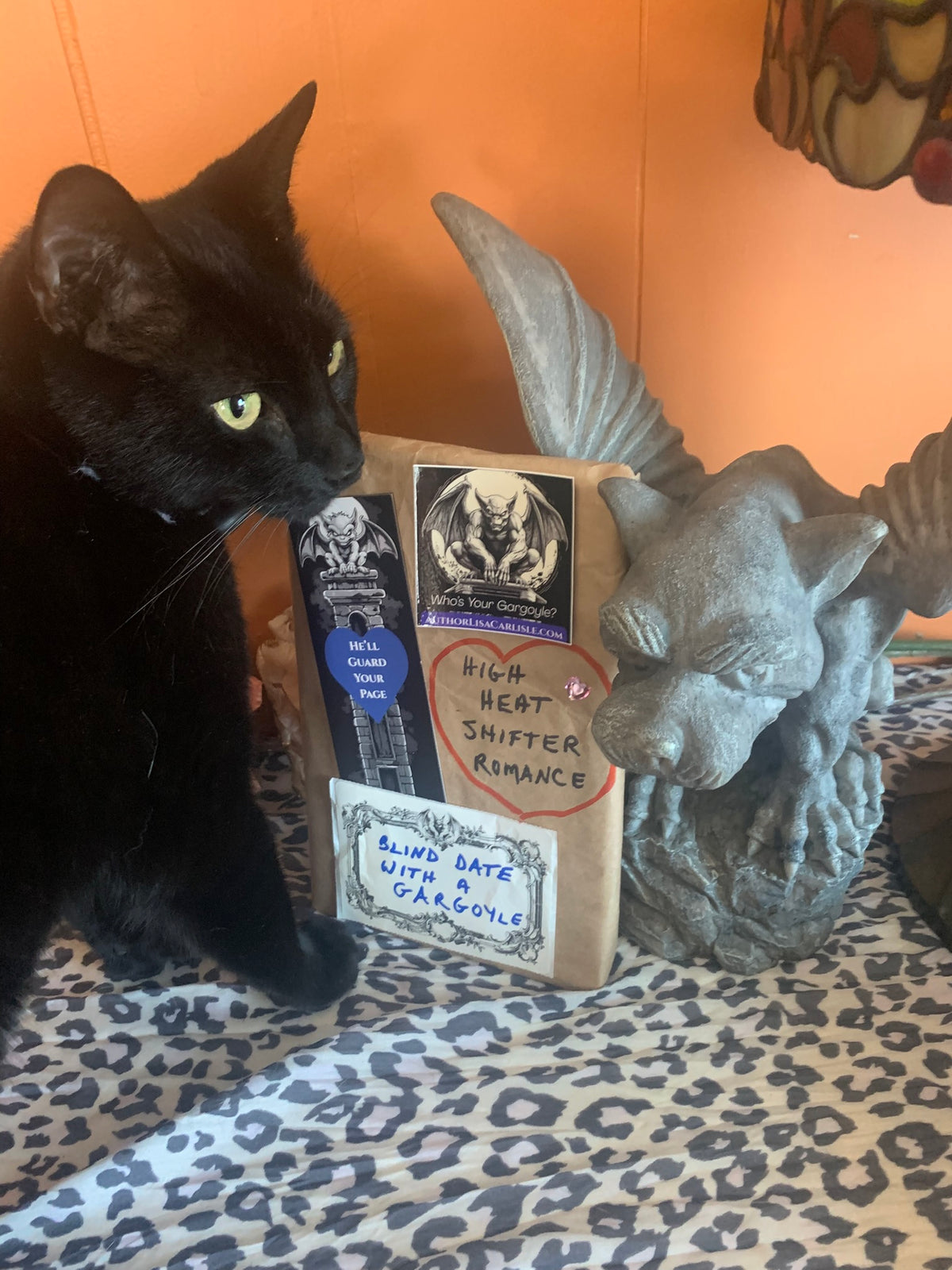 A black cat with yellow eyes sits beside a stack of romance book covers and a gray gargoyle figurine on a leopard-print surface against an orange wall.